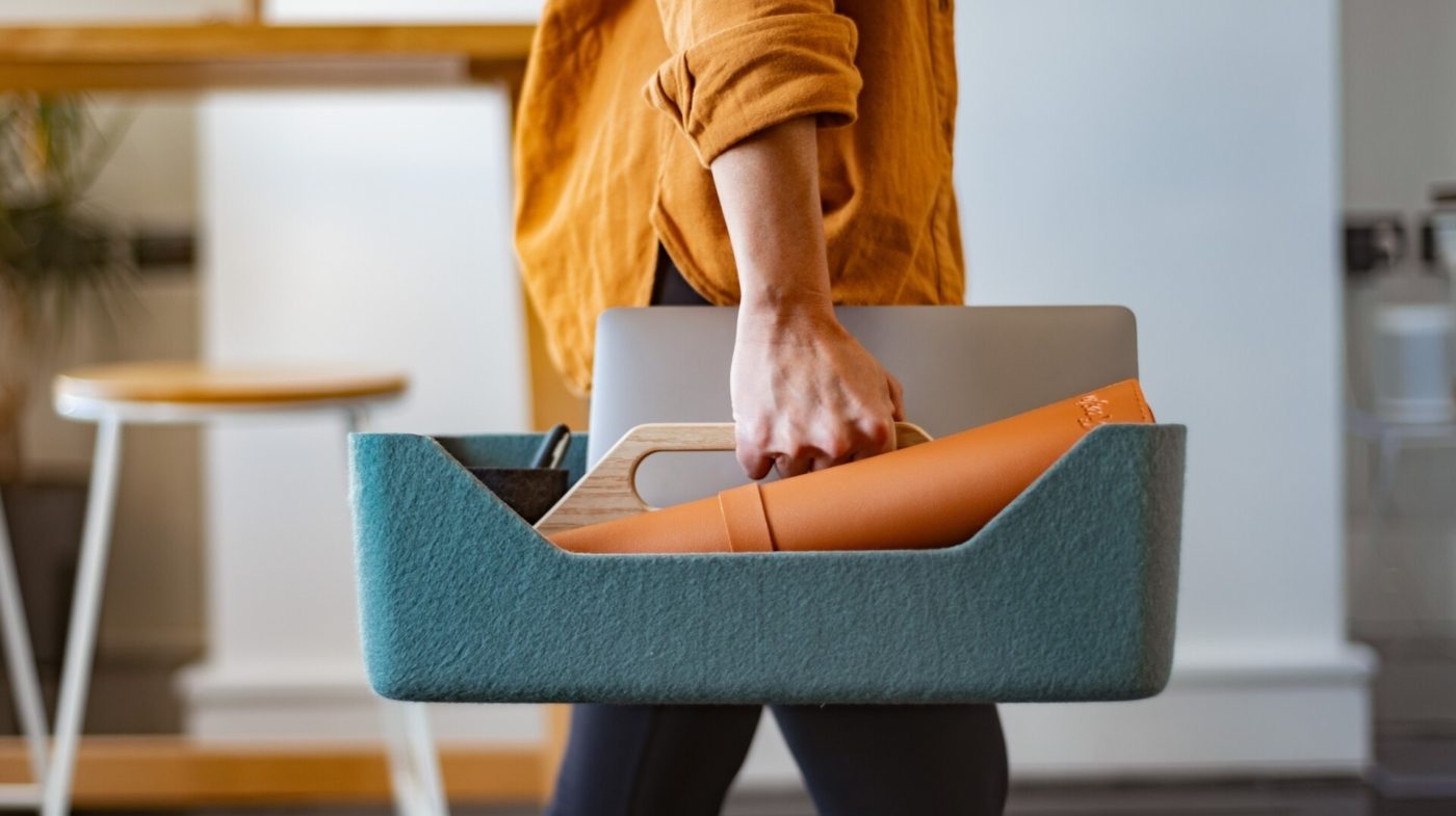 Person carrying a teal desk‑sharing caddy containing a laptop and work items through an office space.