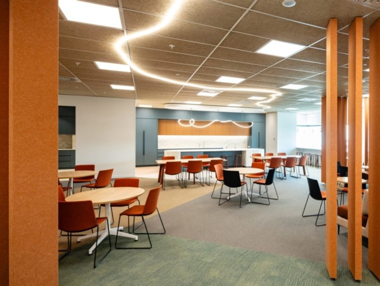 This image shows a modern breakout and dining space within the GACHC office interior. The area features multiple café‑style tables and chairs arranged to support informal meetings, dining, and social interaction. Vertical timber screen elements create partial separation and visual interest while maintaining openness across the space. The interior includes patterned carpet flooring, acoustic ceiling panels, and curved feature lighting that guides movement through the area. The overall design supports flexible use, collaboration, and staff wellbeing within the workplace.